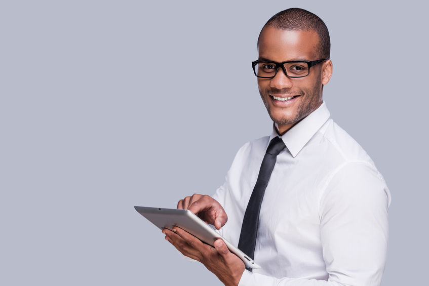 Businessman with digital tablet. Confident young African man in shirt and tie working on digital tablet and smiling while standing against grey background